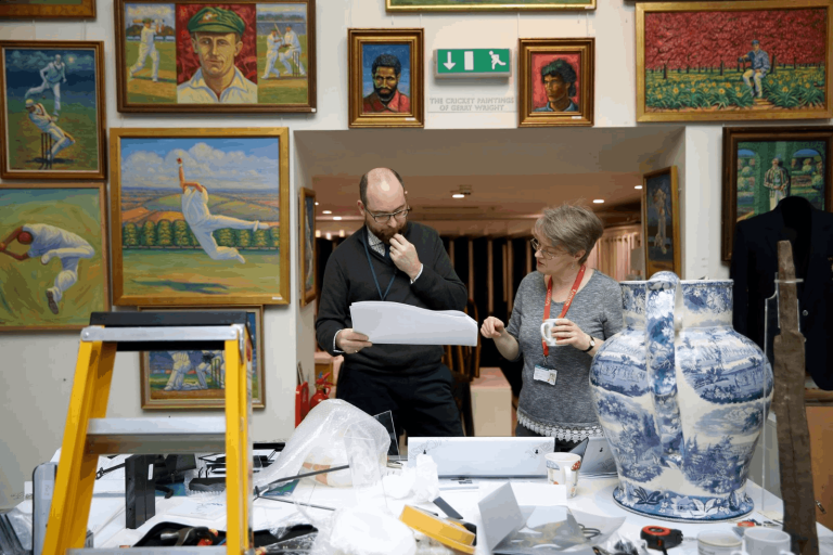 Man and woman looking at a peice of paper. Paintings on wall behind. Large jug and random items in table in foreground.